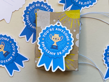 A small, brown kraft gift box decorated with hand-drawn yellow sun and dot designs is topped with a blue ribbon-shaped gift tag with white elastic string that says You’re Awesome and features a golden trophy illustration.