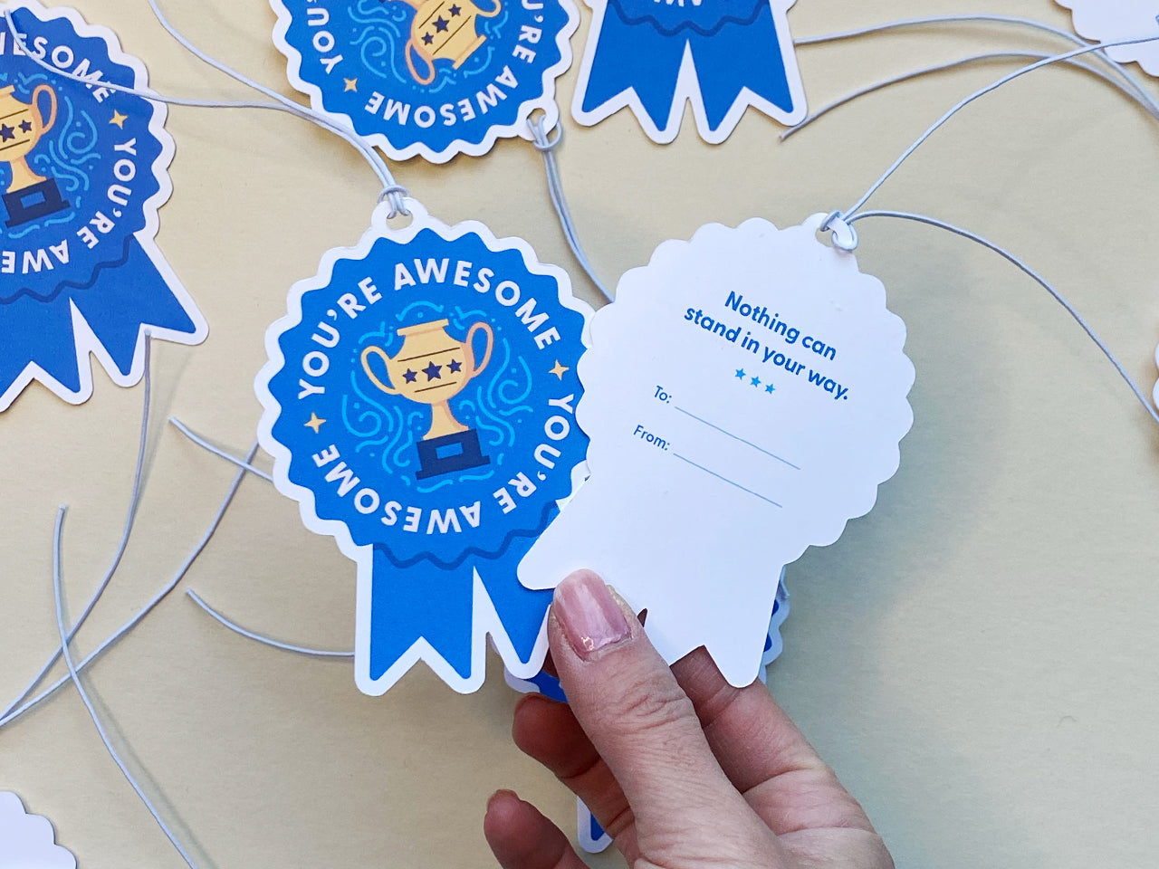 A hand holds a blue ribbon-shaped gift tag, showing the back side with the text Nothing can stand in your way and spaces for To and From. The front side of the tags, featuring You’re Awesome with a golden trophy.