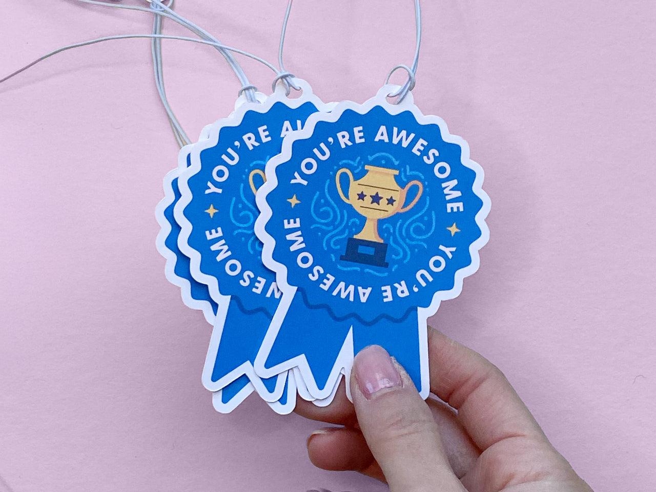 A hand holding two blue ribbon-shaped gift tags that say You’re Awesome with a golden trophy illustration. The tags are tied with white strings and layered together against a pastel pink background, creating a bright display of the celebratory tags.