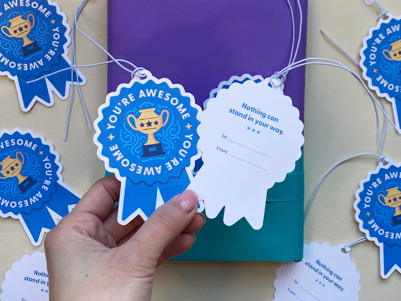 A hand holds a blue ribbon-shaped gift tag showing the back, which reads Nothing can stand in your way with spaces for To and From. The front of the tag, featuring You’re Awesome and a golden trophy design, is visible in the background.