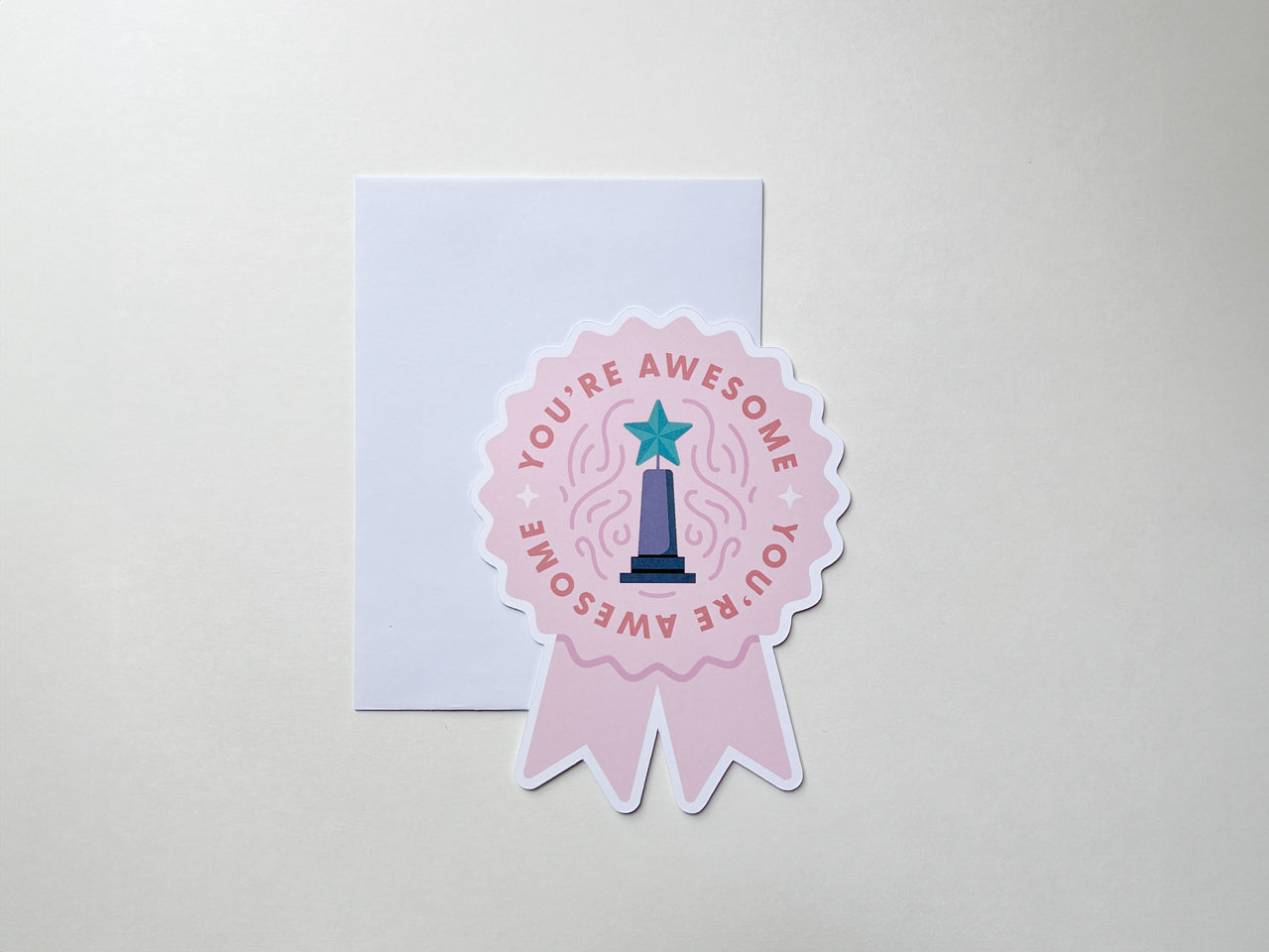 A teal die-cut note card shaped like a prize ribbon features the phrase You're Awesome in bold pink letters, along with an illustration of a trophy with a purple star. Surrounding it are partial views of other ribbon-shaped note cards.