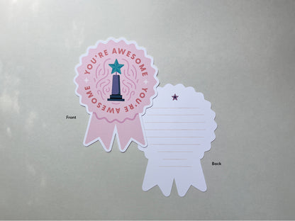 A pink die-cut note card is shown from the front and back. The front reads You're Awesome in burnt orange color with a teal star-topped trophy, while the back has lined space to write in a personal message and a small purple star at the top.