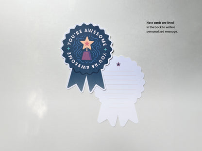 An indigo blue prize ribbon-shaped note card is shown alongside its back side, which is lined for writing. The card reads You're Awesome with a yellow star-topped trophy illustration, and the back of note includes lines to write a personal message.