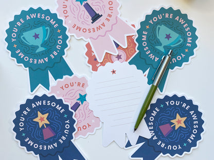 A collection of die-cut note cards shaped like prize ribbons is laid out on a white surface. Each card displays the phrase You&#39;re Awesome with illustrations of trophies. One card is flipped to its back, lined for writing a personal message.