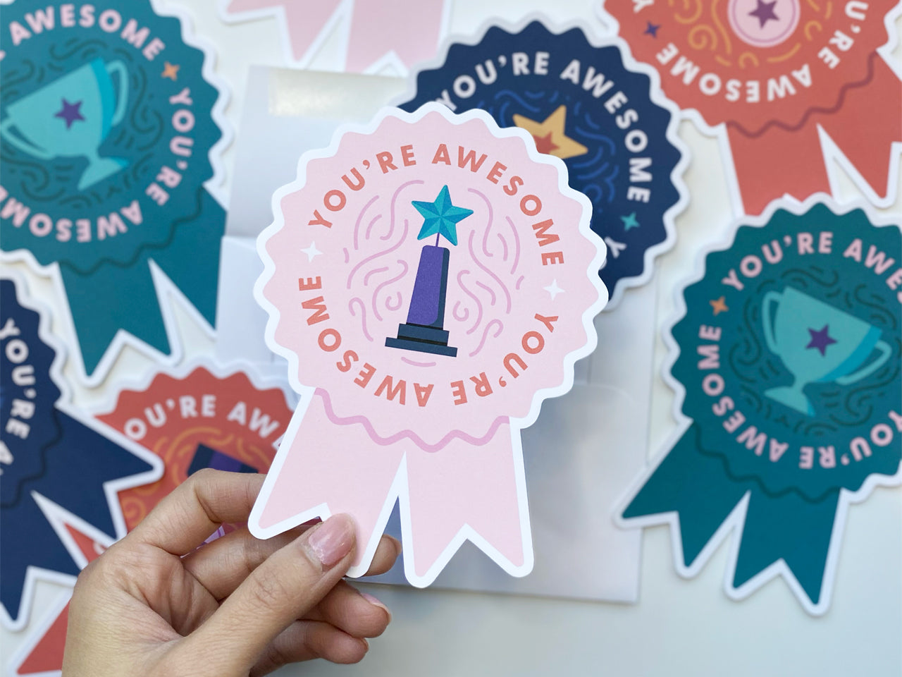 A hand holds a pink die-cut note card shaped like a prize ribbon, with You're Awesome written in bold letters and an illustration of a teal star-topped trophy. In the background, several other ribbon-shaped note cards.