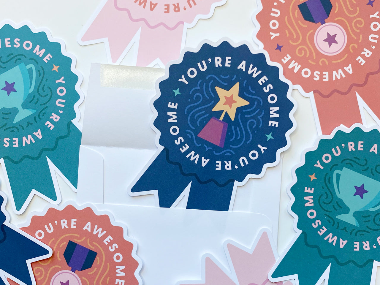 A collection of colorful die-cut note cards shaped like prize ribbons is spread across a white surface. The cards feature You're Awesome in bold letters with trophy illustrations in teal, pink, orange, and indigo.