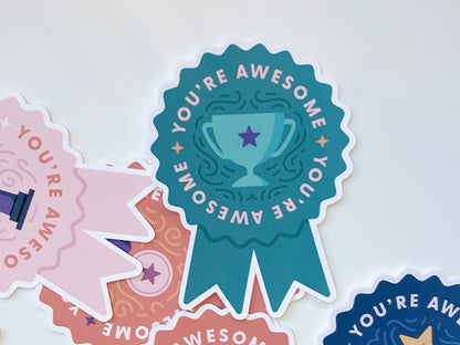 A teal die-cut note card shaped like a prize ribbon features the phrase You're Awesome in bold pink letters, along with an illustration of a trophy with a purple star. Surrounding it are partial views of other ribbon-shaped note cards.