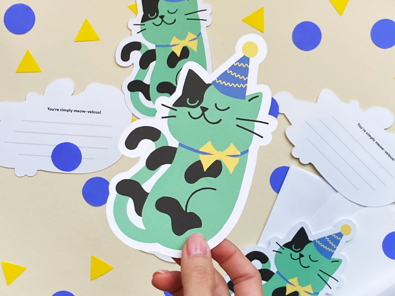 A hand holding a large, cat-shaped note card featuring a playful green cat illustration. The cat wears a blue party hat with a yellow pompom and a matching yellow bowtie. The back of the note card is lined for writing personalized messages.