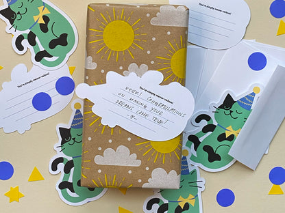 A gift wrapped in brown kraft paper decorated with bright yellow suns and white fluffy clouds. A green party-cat note card is attached as a gift tag, adding a playful and festive touch. Also shown is the back lined note cards for handwritten message.