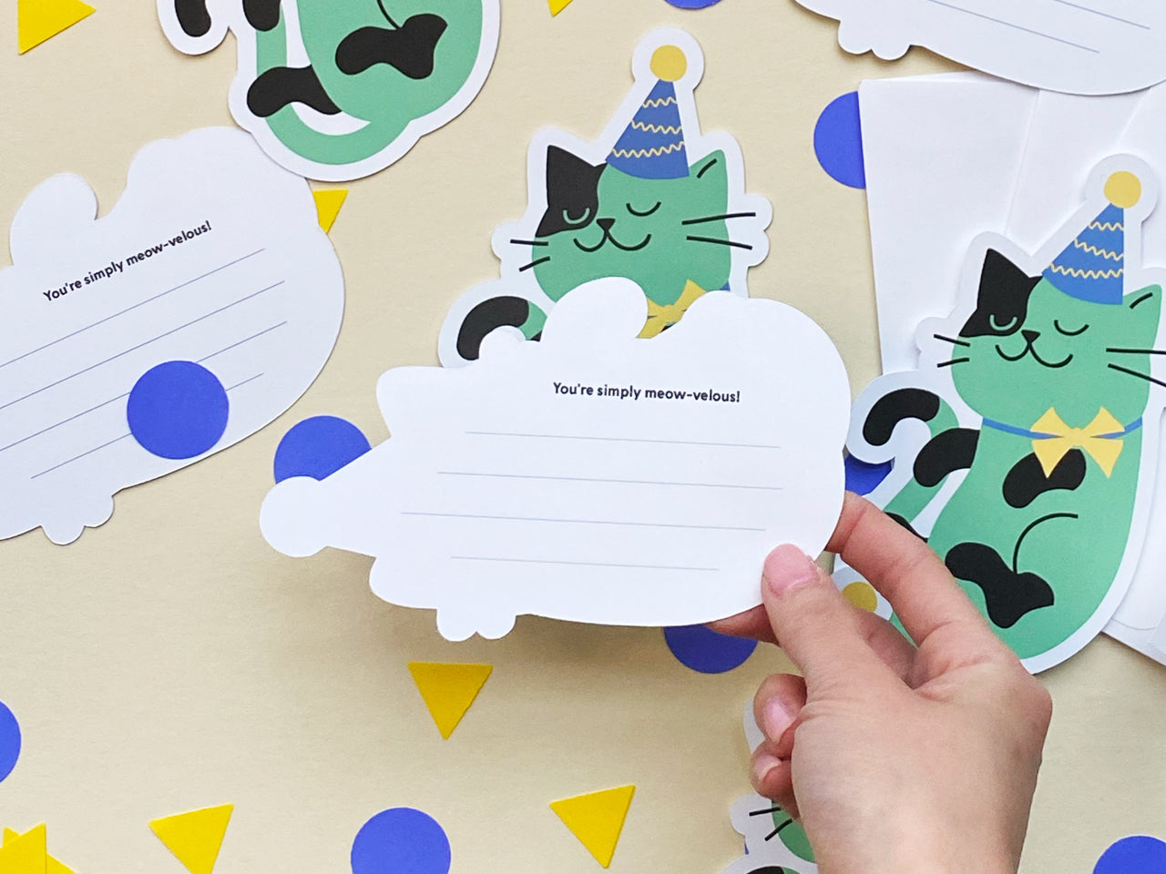 A hand holding the back of a lined note card with a message You’re simply meow-velous! printed at the top. The card is lined for writing and surrounded by cat-shaped note cards featuring a green cat in a party hat and yellow bowtie.