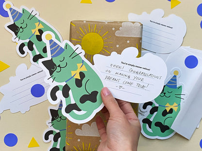 A hand holds a die cut note card with a handwritten congratulatory message. A gift wrapped in sunny kraft paper sits among cat-themed stationery, including green party-cat wearing purple party hat and yellow bowtie and envelopes.