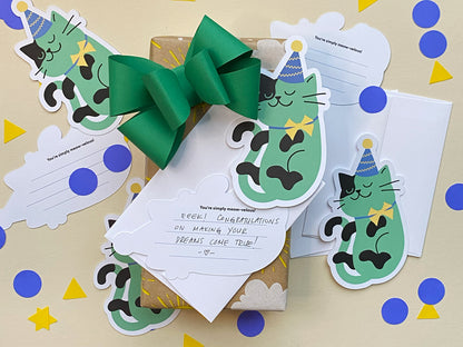 A gift wrapped in kraft paper with yellow sun and cloud designs, topped with a large green bow. Surrounding the gift are party cat note cards featuring a green cat with a purple hat and yellow bowtie, along with the lined back shown.