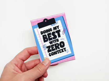 Hand holding a rectangular magnet with a pink grid background and a blue clipboard illustration holding lined paper. Bold black text reads “Doing My Best With Zero Context.”